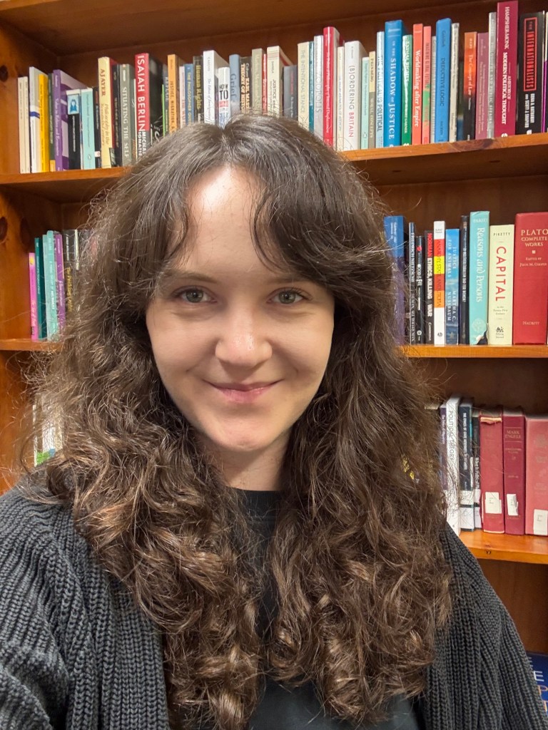 A selfie of Eilidh standing in front of a bookcase. She has brown hair and is wearing a black t-shirt and black cardigan.